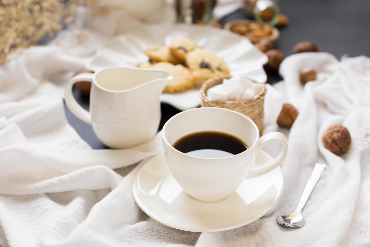 Tasse Kaffee, Untertasse und Sahneserver neben Gebäck auf zerknitterter Tischdecke foto