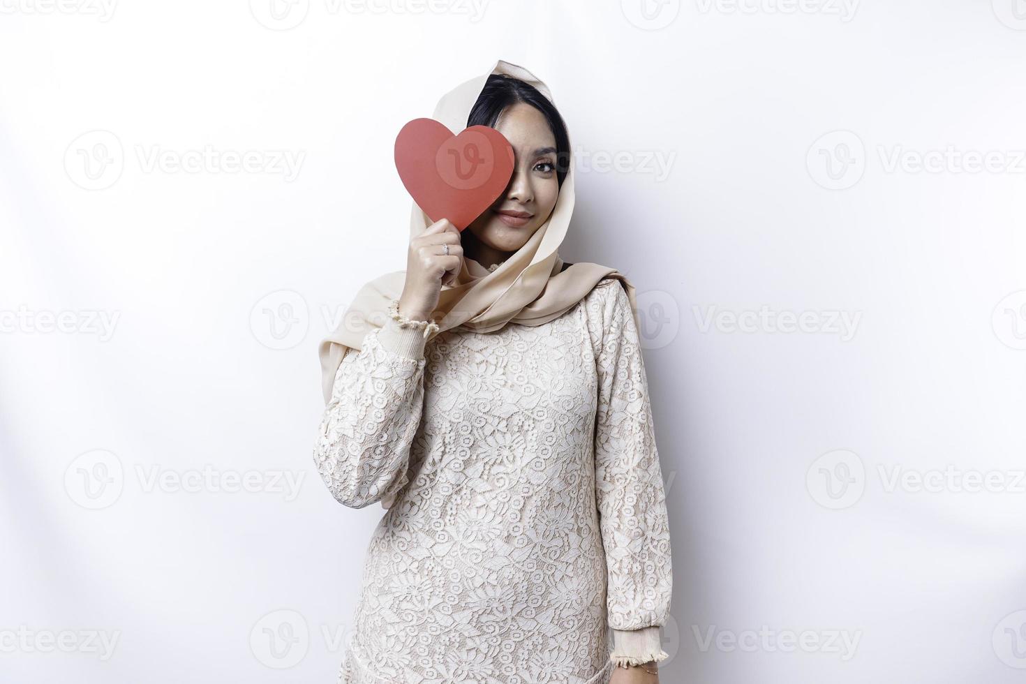 ein glücklich jung asiatisch Muslim Frau tragen ein Hijab fühlt sich romantisch Formen Herz Geste ausdrücken zärtlich Gefühle und halten ein rot herzförmig Papier foto