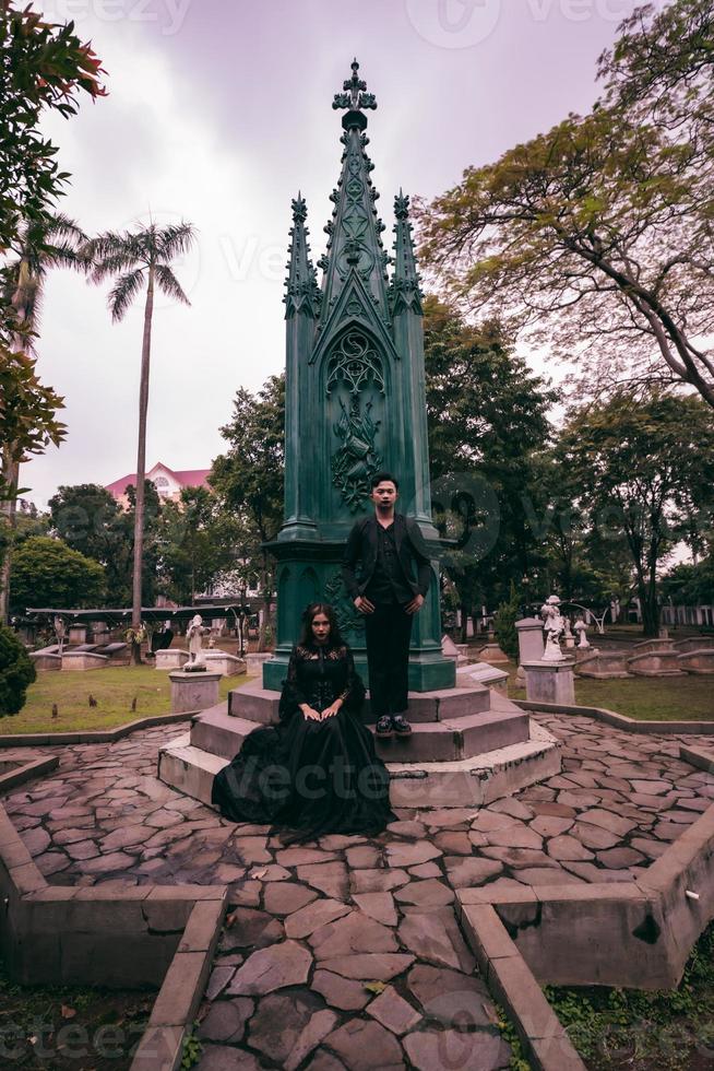 ein Paar von Liebhaber Sitzung und Stehen im Vorderseite von ein Beerdigung Monument im alles schwarz Kleider und unheimlich Ausdrücke auf Halloween foto