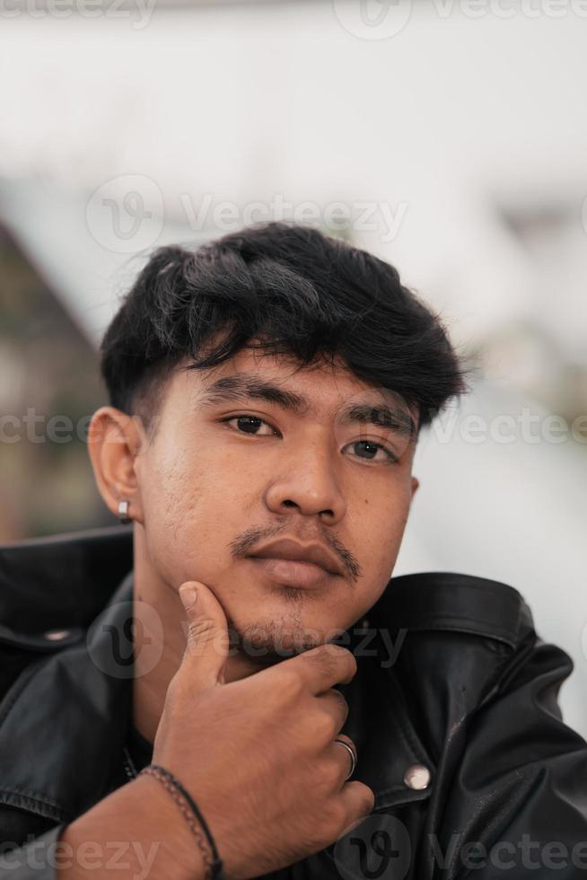 ein asiatisch Mann mit ein frech Gesicht tragen ein Kette Halskette und ein schwarz Leder Jacke im ein Cafe foto