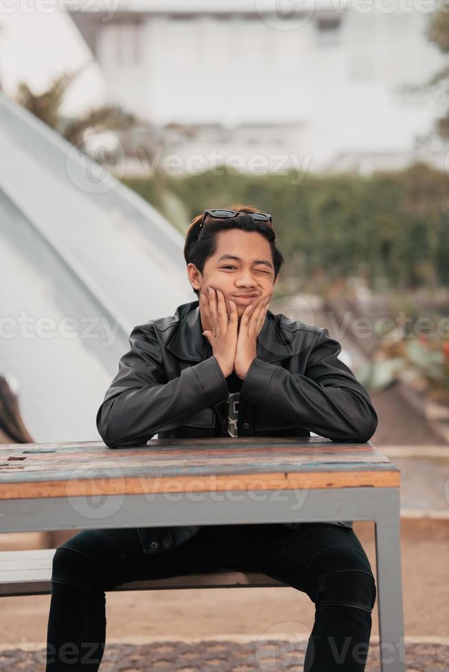 ein asiatisch Mann mit ein mollig Gesicht tragen Sonnenbrille und ein schwarz Leder Jacke während Sitzung beim ein Cafe Tabelle foto