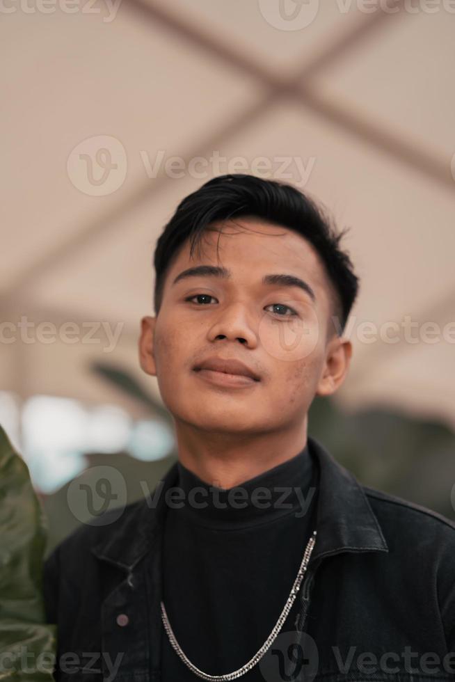 ein asiatisch Mann im ein schwarz Denim Jacke posieren mit ein heiter Gesicht und sehr gut aussehend im ein Cafe foto