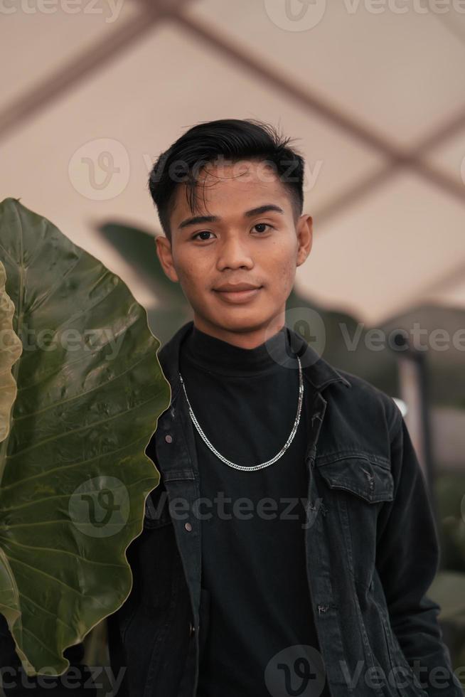 ein asiatisch Junge im ein schwarz Denim Jacke posieren mögen ein frech Junge mit ein Kette um seine Hals im ein Park foto