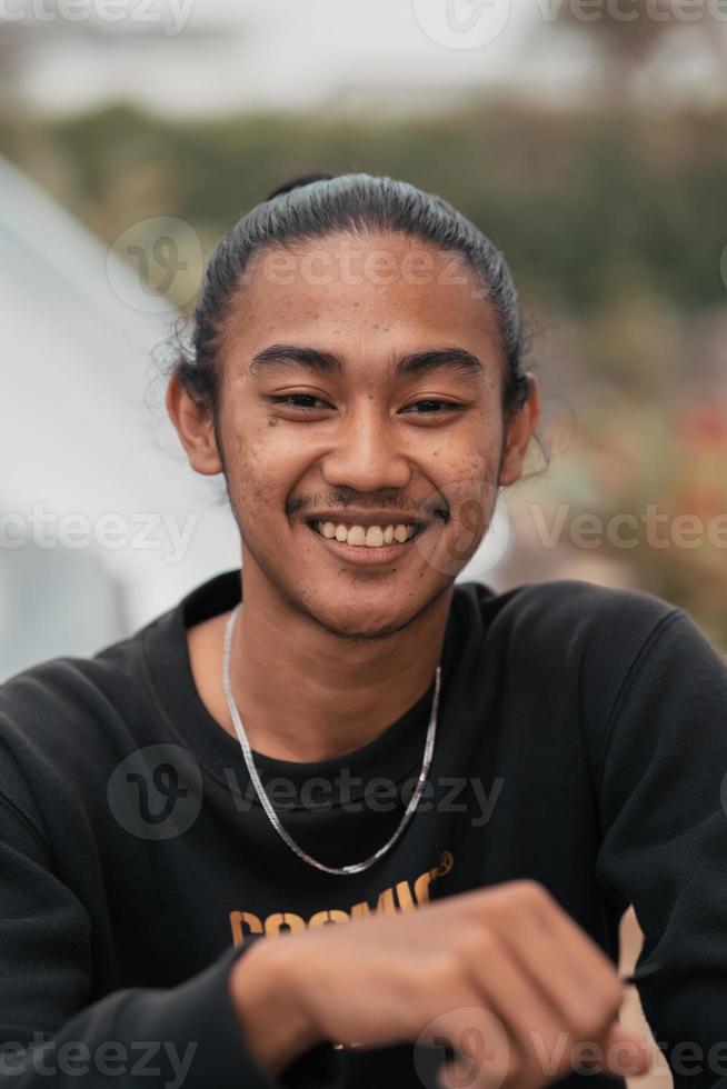 ein asiatisch Mann mit seine Haar gebunden oben und tragen ein schwarz Sweatshirt Jacke ist Sitzung entspannend beim ein Cafe Tabelle wann Treffen seine freunde foto