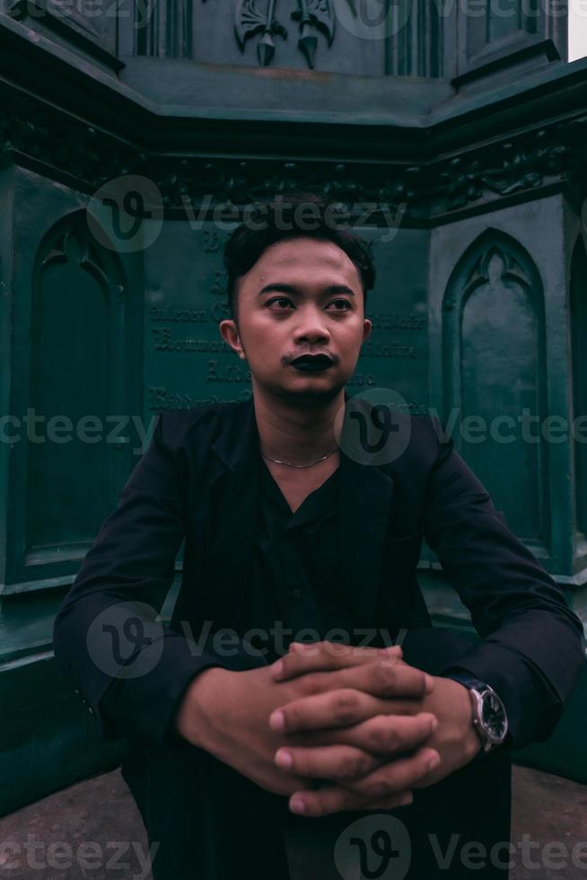 ein asiatisch Mann im ein schwarz passen ist Sitzung allein im Vorderseite von das Beerdigung Monument mit ein sehr unheimlich Ausdruck foto