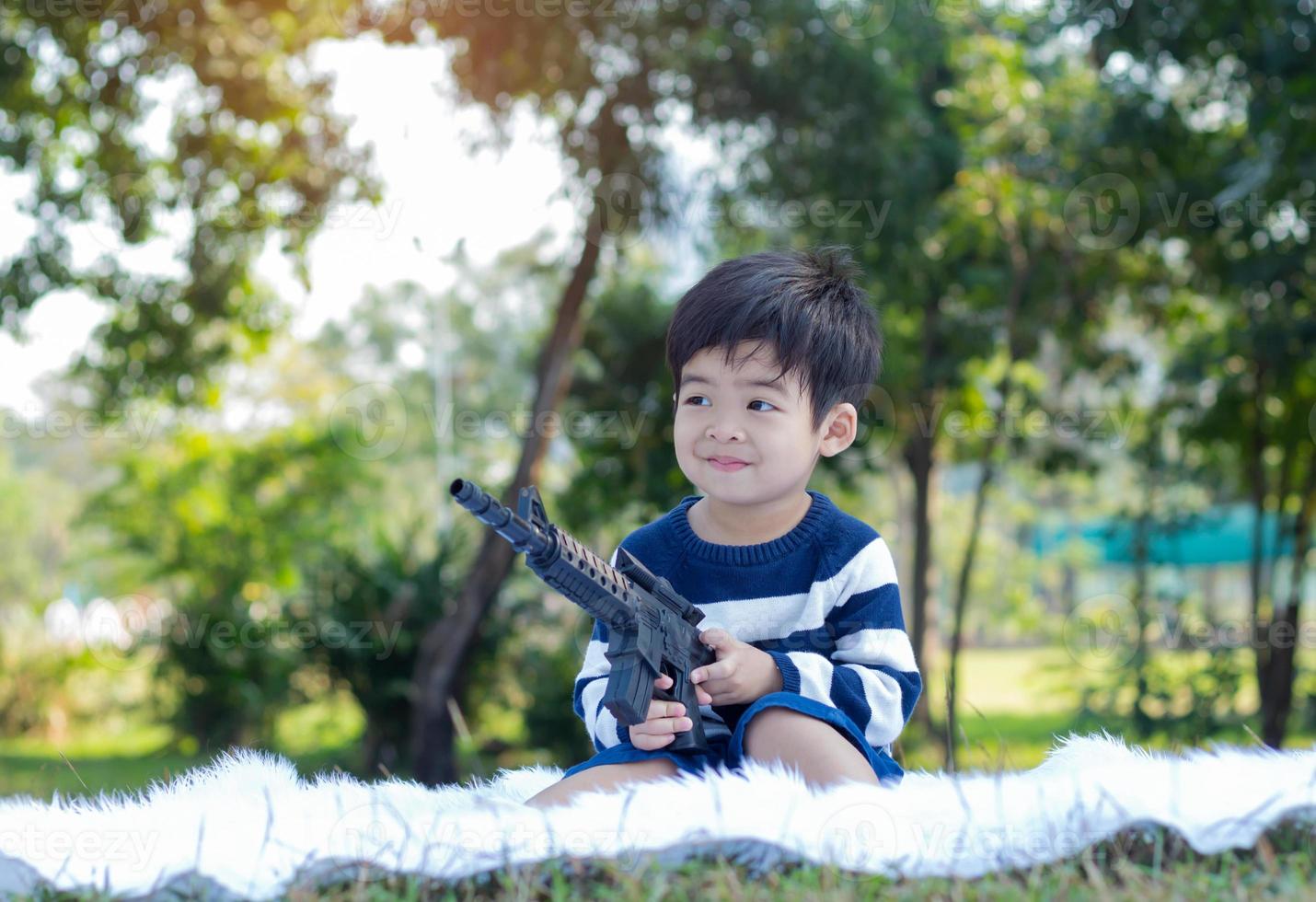 asiatischer Junge, der in einem Park an einem klaren Morgen sitzt und eine Spielzeugpistole hält foto