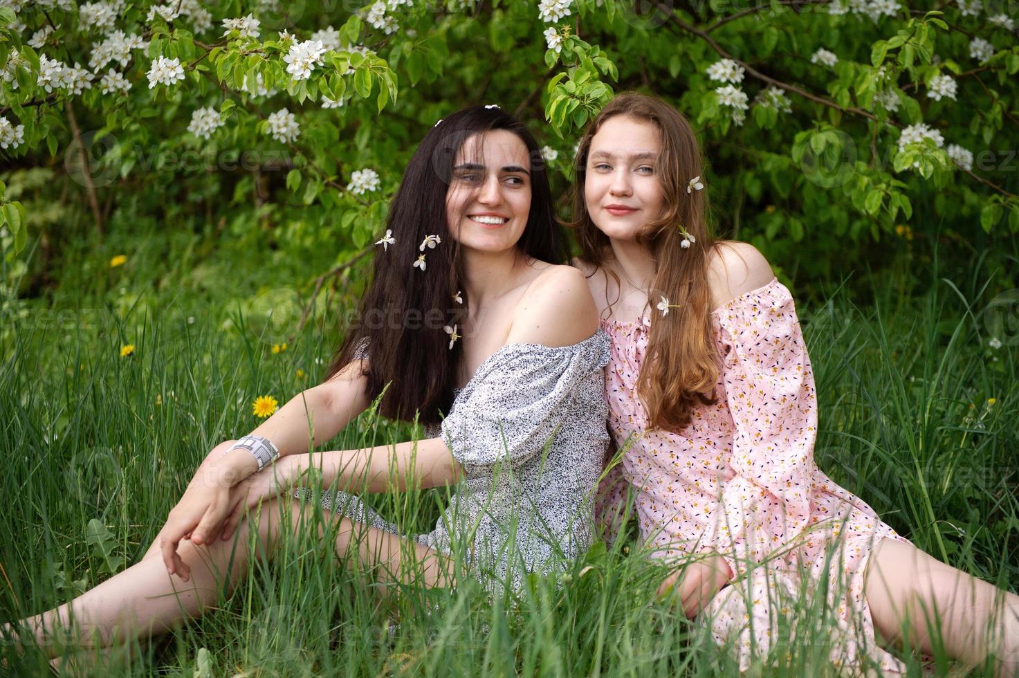 zwei jung Mädchen im Kleider sind Sitzung auf das Grün Gras unter ein Weiß Baum und Lachen foto
