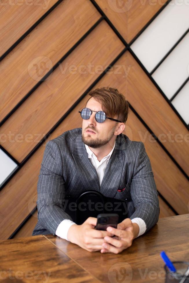 ein jung Mann im ein passen und Sonnenbrille ist Sitzung beim ein Büro Schreibtisch mit ein Telefon foto