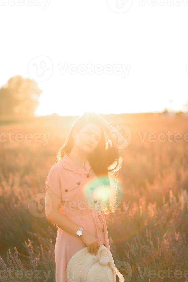 ein heiter jung Mädchen im ein Rosa Kleid und ein Stroh Hut im ihr Hände steht unter Lavendel Gebüsch. Sonnenuntergang. foto