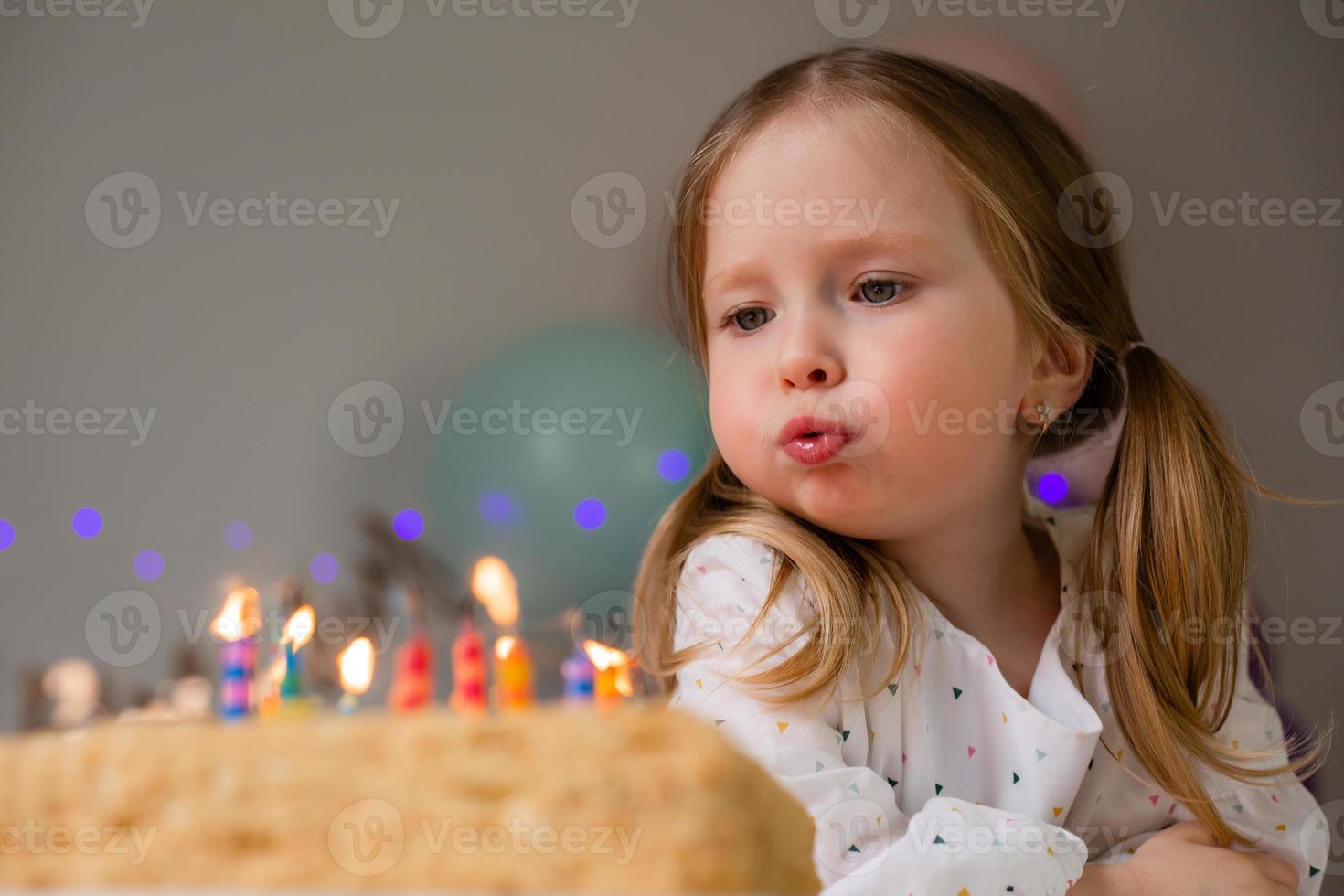 süß wenig Mädchen weht aus Kerzen auf ein Geburtstag Kuchen beim Zuhause gegen ein Hintergrund von Luftballons. Kinder Geburtstag foto