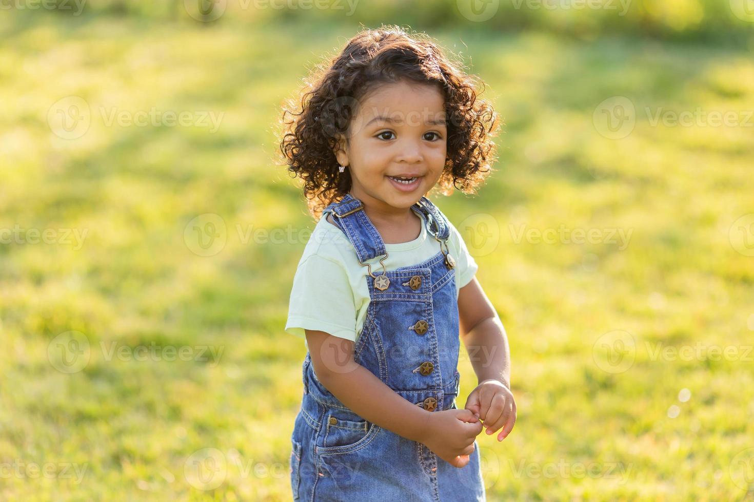 Porträt wenig glücklich spielerisch Kleinkind dunkel Mädchen im ein Denim Sommerkleid Stehen im das Garten auf ein sonnig Tag. Gehen im das frisch Luft. Konzept von ein glücklich Kindheit. Raum zum Text. hoch Qualität Foto