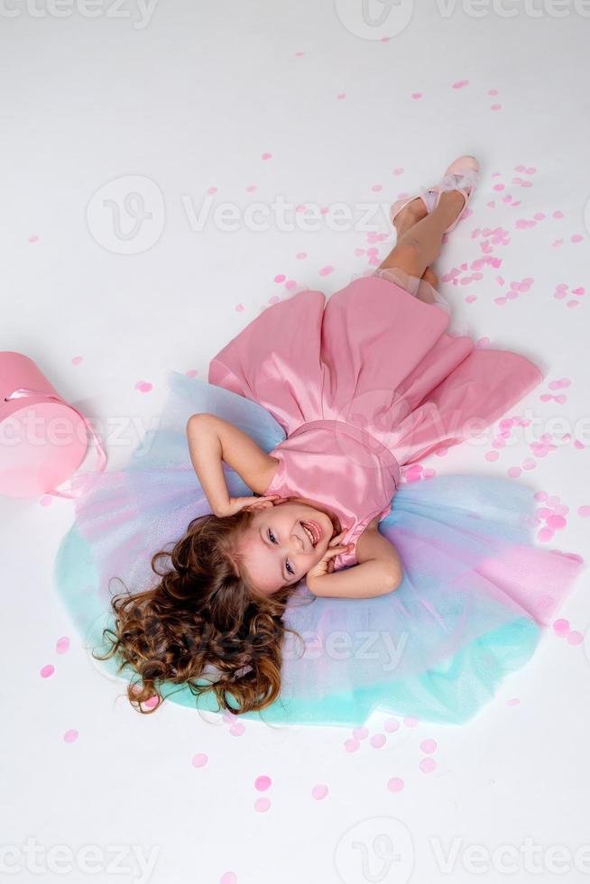 schön wenig Mädchen im ein schick Rosa Kleid Lügen auf das Fußboden übersät mit Konfetti. oben Sicht. Mode und Stil. Kind feiert seine Geburtstag. Foto im das Studio. Raum zum Text. hoch Qualität Foto