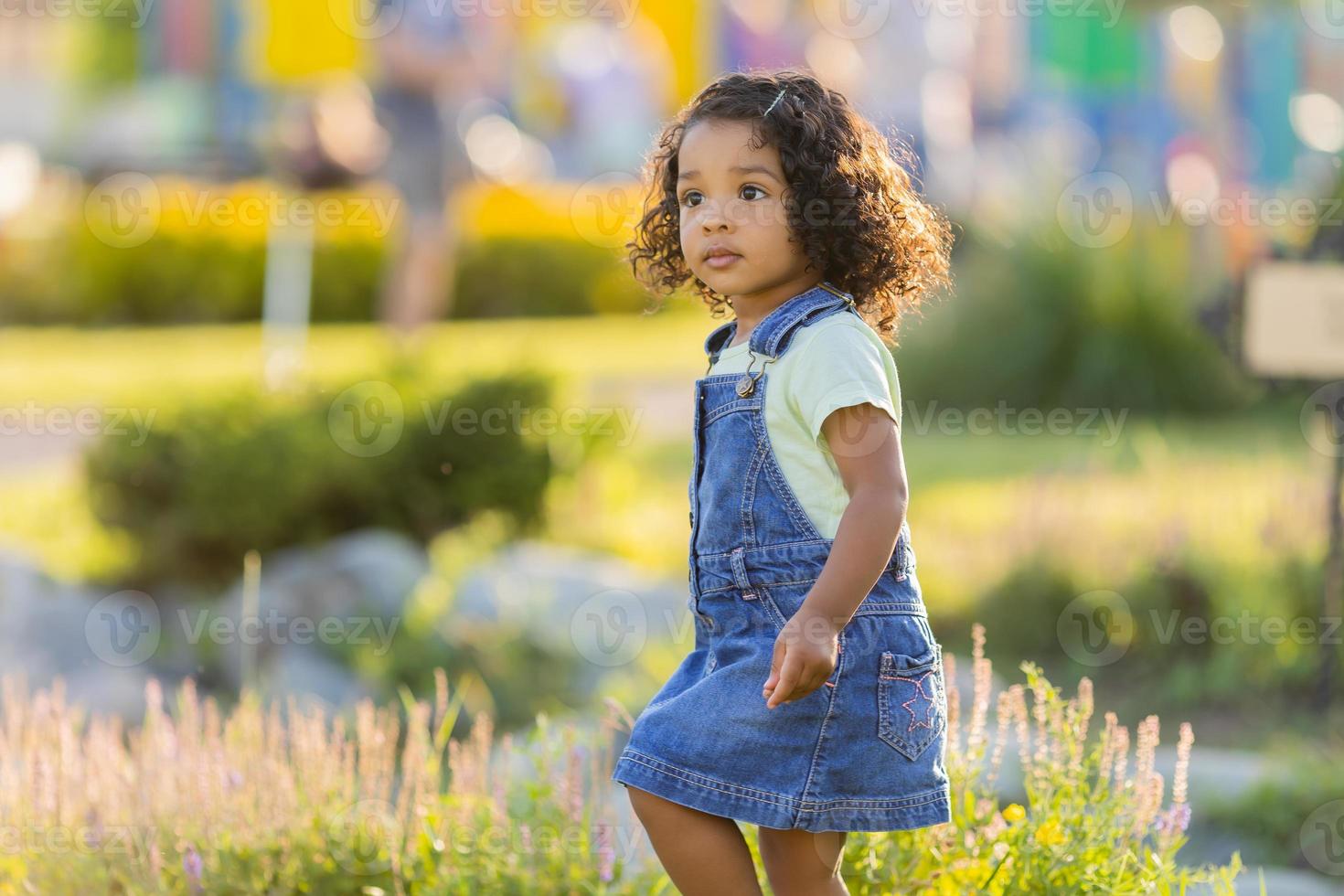 Porträt wenig glücklich spielerisch Kleinkind dunkel Mädchen im ein Denim Sommerkleid Stehen im das Garten auf ein sonnig Tag. Gehen im das frisch Luft. Konzept von ein glücklich Kindheit. Raum zum Text. hoch Qualität Foto