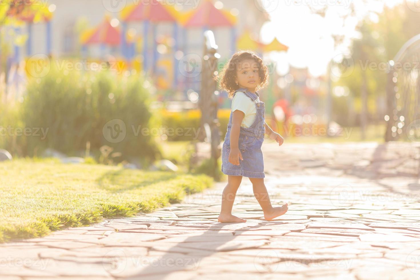 Porträt wenig glücklich spielerisch Kleinkind dunkel Mädchen im ein Denim Sommerkleid Stehen im das Garten auf ein sonnig Tag. Gehen im das frisch Luft. Konzept von ein glücklich Kindheit. Raum zum Text. hoch Qualität Foto