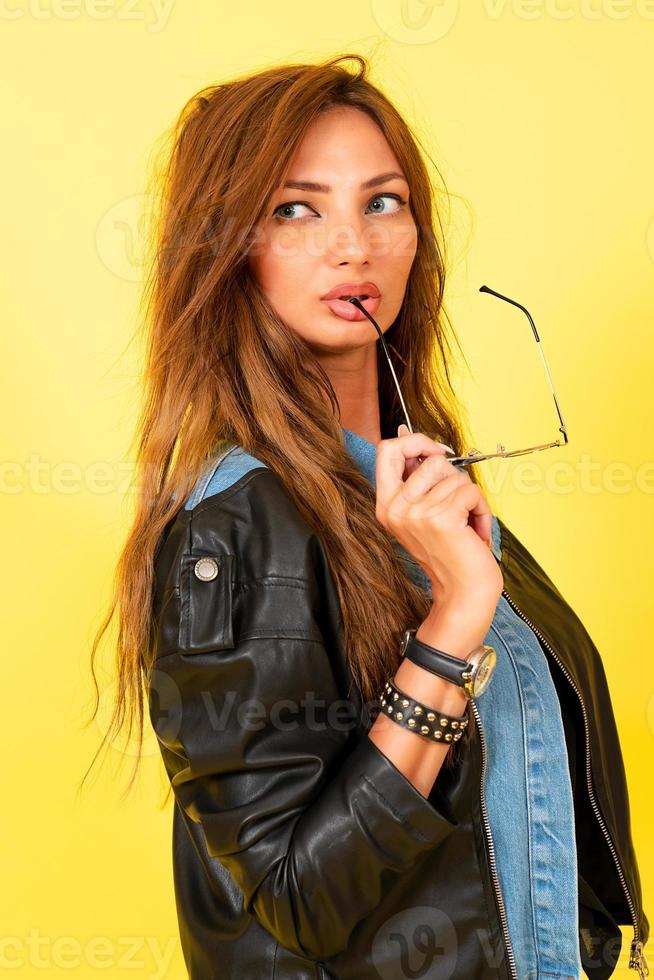 Gelb Hintergrund, ein Mädchen im ein Leder Jacke im das Stil von Felsen, zeigt an Emotionen, hält Brille im ihr Zähne foto