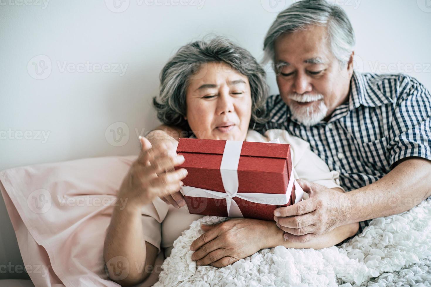 lächelnder älterer Ehemann, der Überraschung macht und seiner Frau im Schlafzimmer Geschenkbox gibt foto