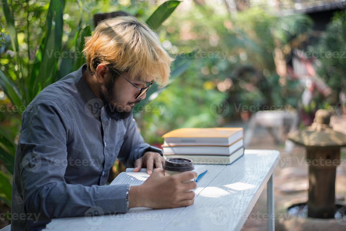 junger Hipster-Bartmann, der Kaffee trinkt, während Bücher im Hausgarten mit Natur liest foto