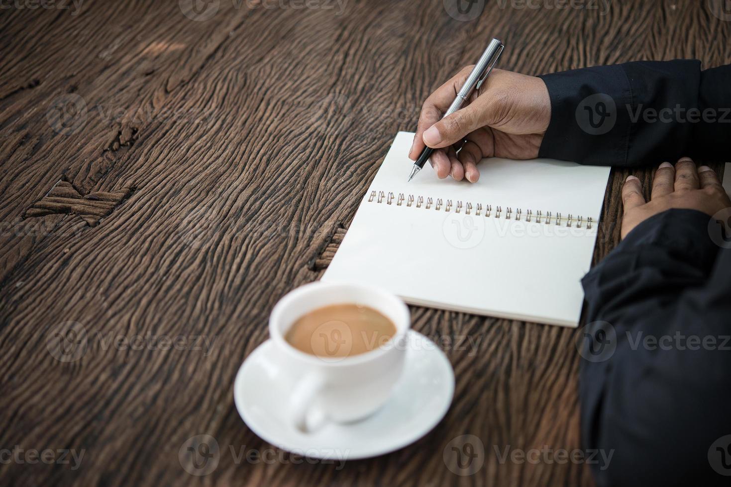 junger Mann, der auf einem Notizbuch schreibt, das an einem rustikalen Holztisch arbeitet foto