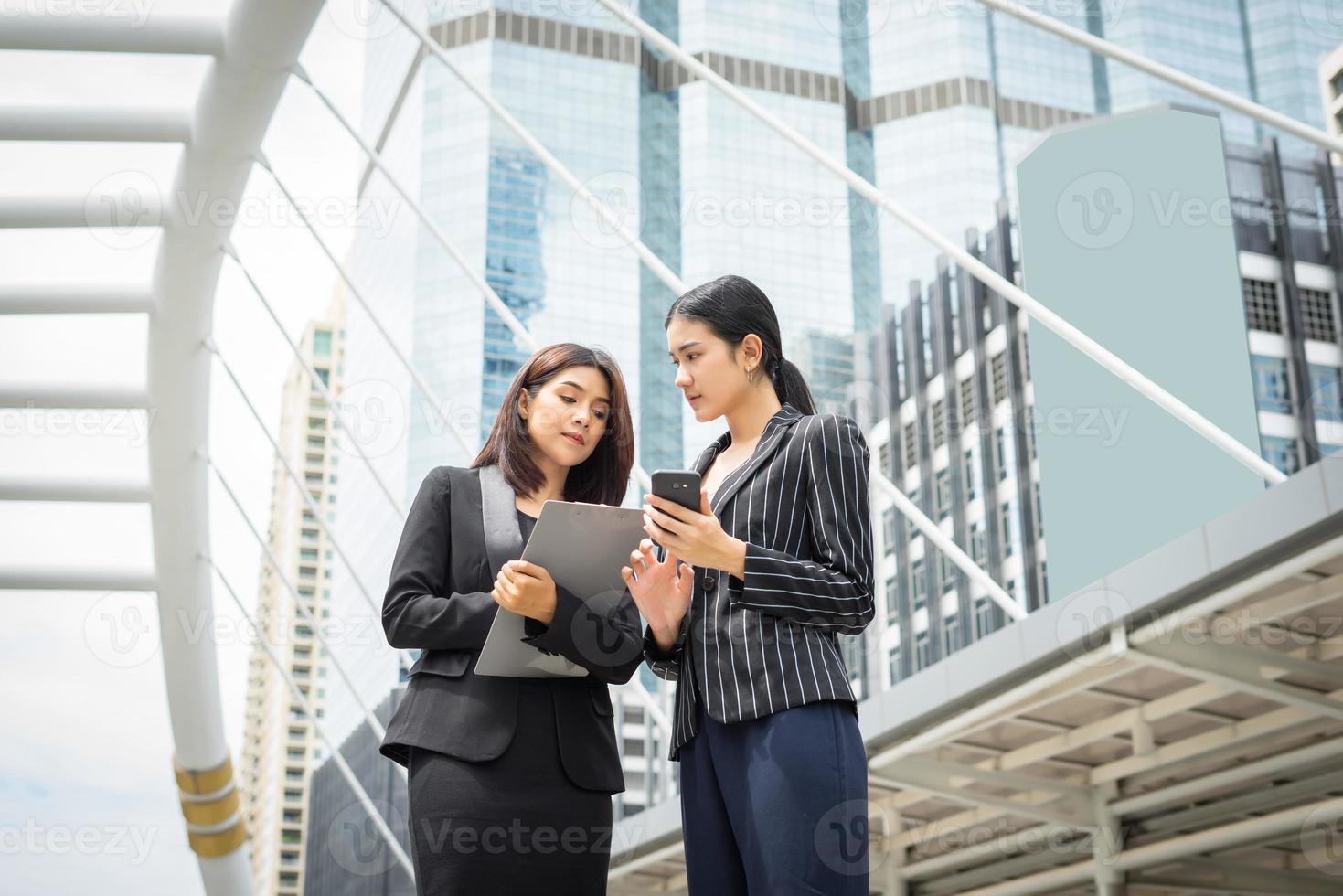 zwei Geschäftsfrau, die ein Smartphone benutzt und Arbeit vor dem Büro bespricht foto