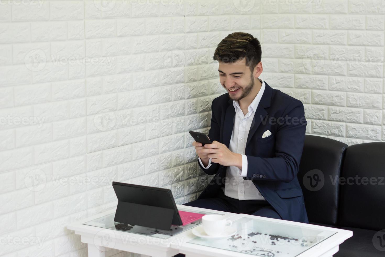 junger Geschäftsmann, der Smartphone während der Arbeit an seinem Laptop im Büro verwendet foto