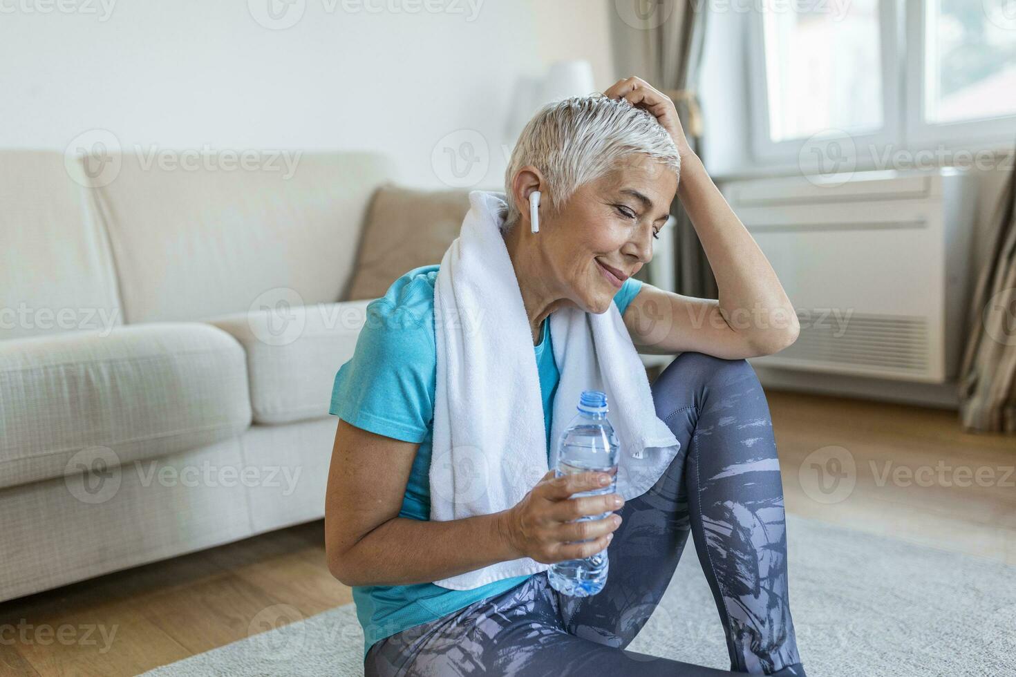 Senior Frau halten Plastik Flasche von Wasser, abwischen Schweiß mit ein Handtuch, erschöpft nach das Täglich Ausbildung. Alten Frau nehmen ein brechen während ausüben beim heim. Sitzung auf Fitness Matte und ruhen foto