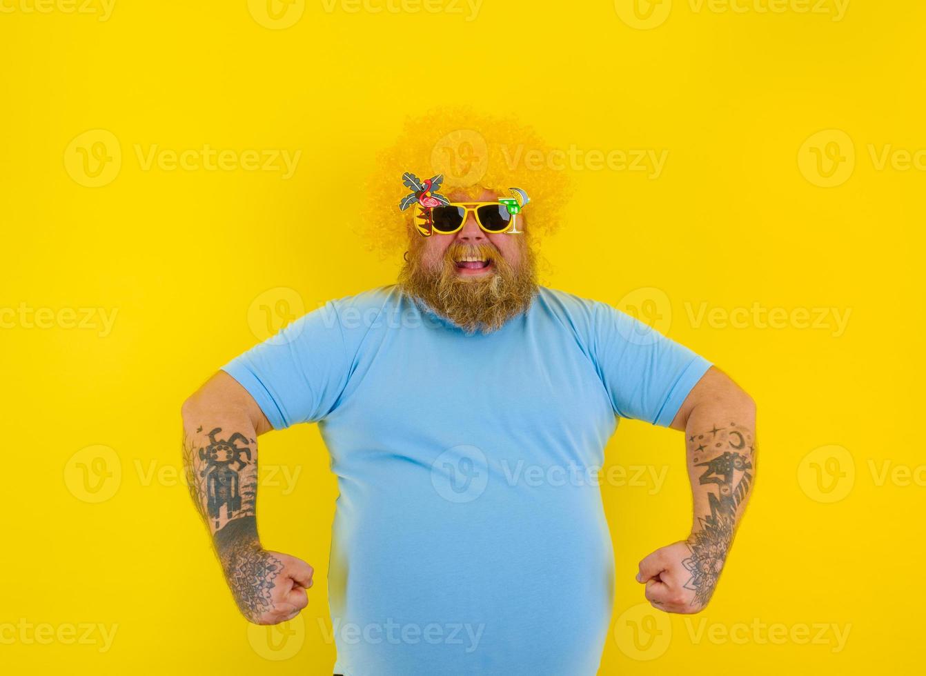 Fett Mann mit Perücke im Kopf und Sonnenbrille zeigt an seine Muskel foto