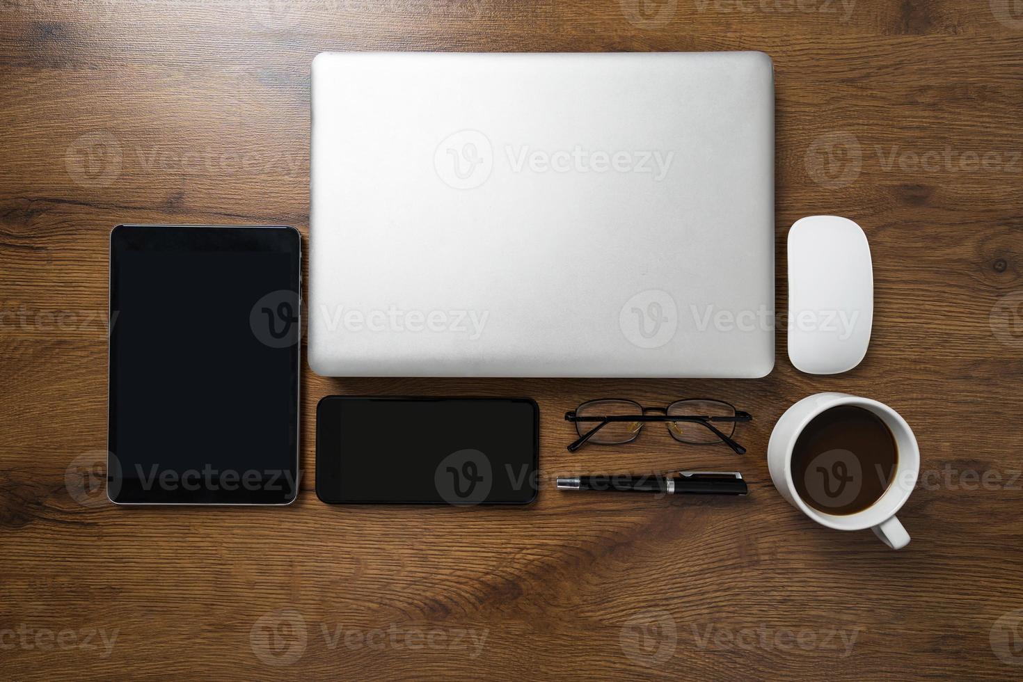 Draufsicht des Arbeitsbereichs mit Laptop, Smartphone, Tablet, Kaffeetasse, Gläsern und Stift auf Holztisch foto