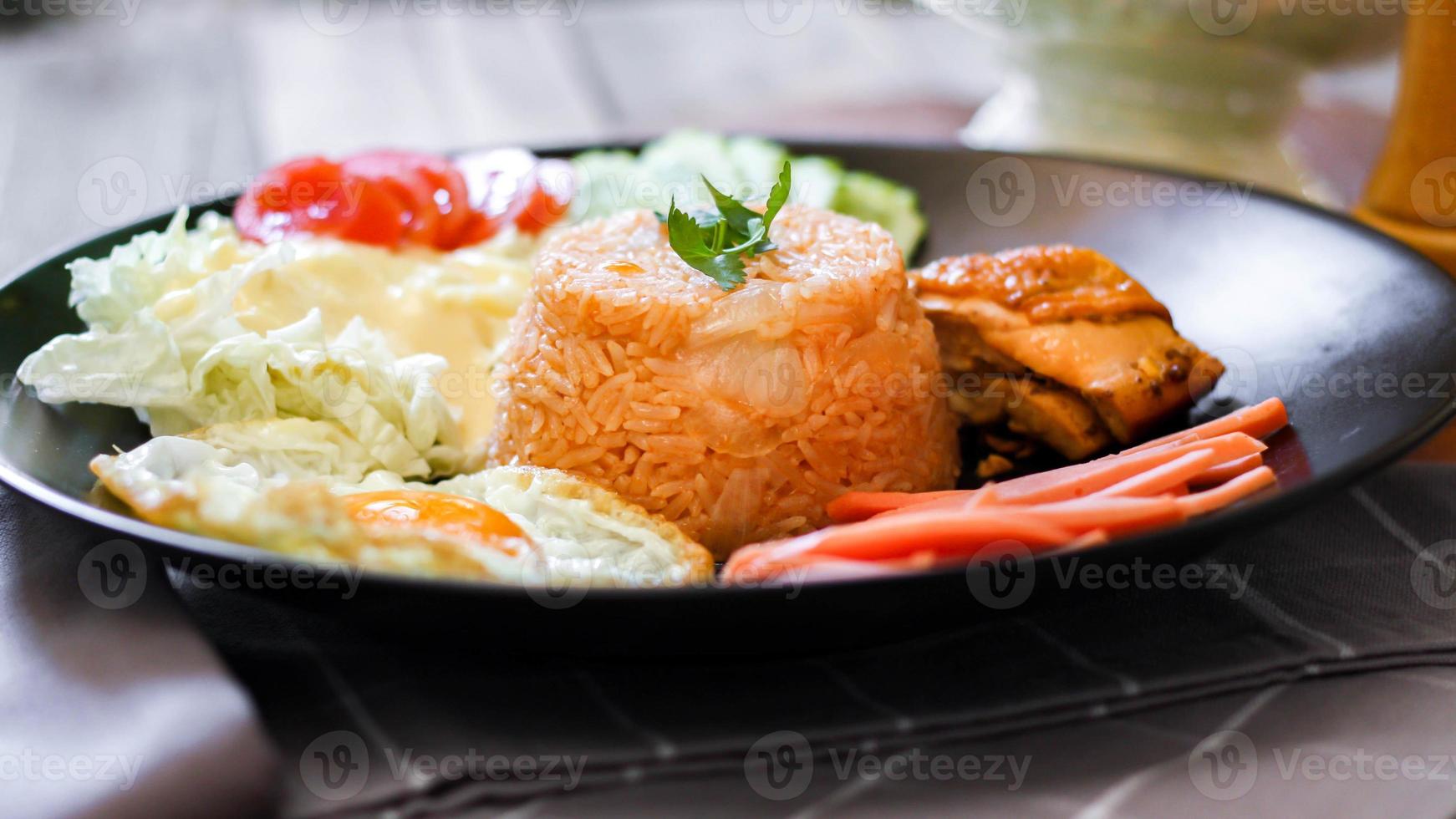 gebraten Reis, gebraten Ei, Gurke, geschnitten Tomate, Grün Salat und gebraten Hähnchen platziert auf ein schwarz Teller wie ein Garnierung. foto