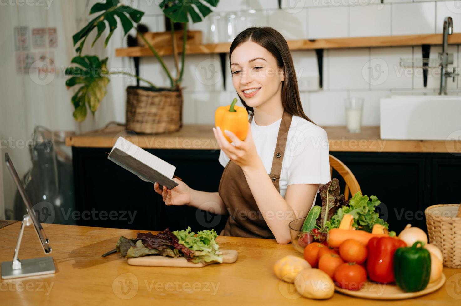 jung schön Frau im das Küche im ein Schürze, frisch Gemüse auf das Tisch, schreibt Nieder ihr Liebling Rezepte, kommt oben mit Ideen zum Geschirr foto