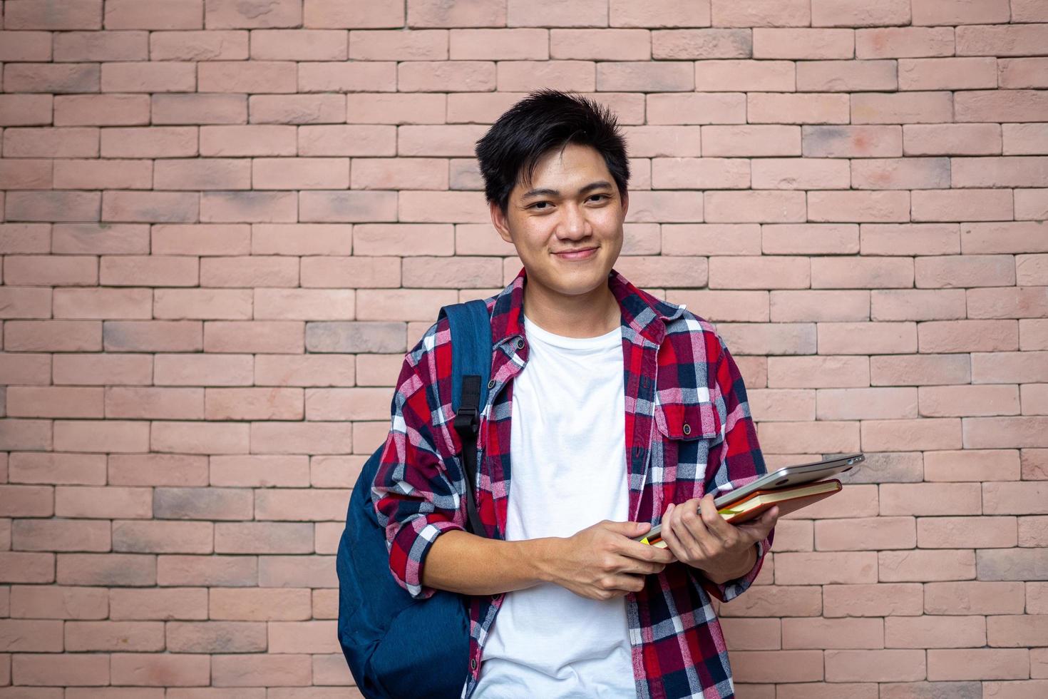 asiatisch männlich Studenten tragen Plaid Hemden. Stehen Nächster zu ein Backstein Wand, Tragen ein Rucksack, Tragen Bücher, Schule Lieferungen, vorbereiten zum lernen, lächelnd. foto