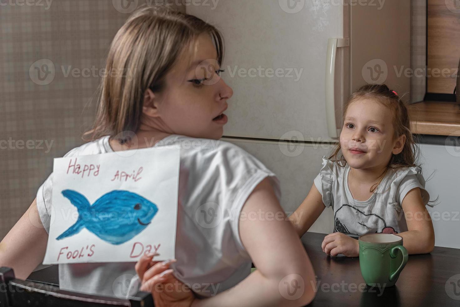 heiter wenig Mädchen geklebt ein Stück von Papier Fisch zu ihr Mutter auf ihr zurück beim Zuhause im das Küche. komisch Familie Witze, April Narren Tag Feier foto