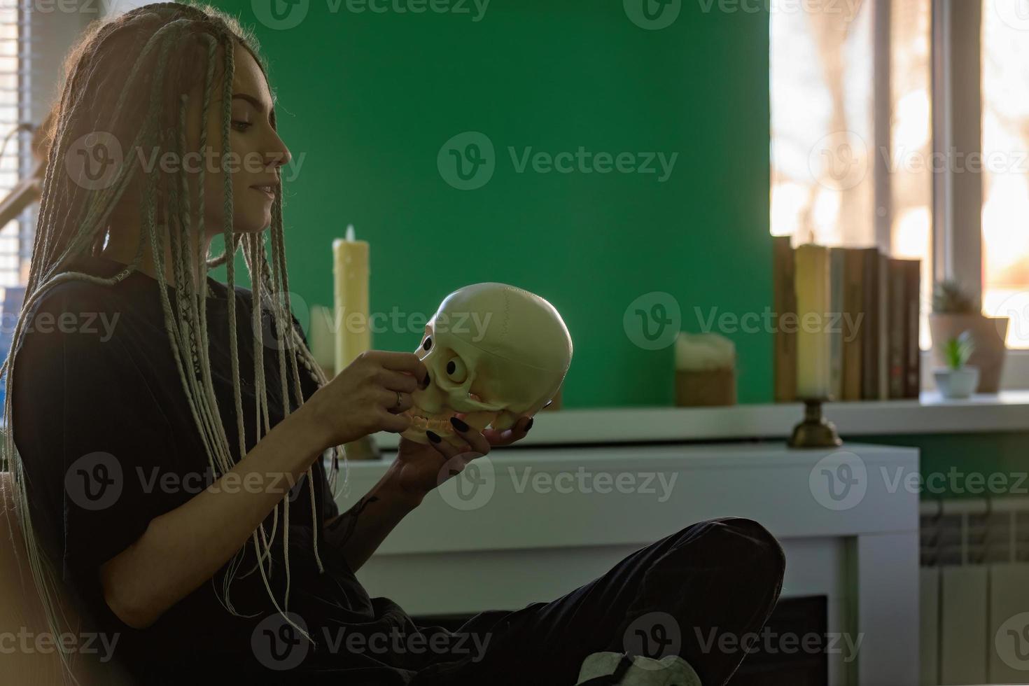 ein jung hell Mädchen im dunkel Kleidung, mit Dreadlocks auf ihr Kopf, hält ein Schädel im ihr Hände. Generation z, gotisch Stil foto