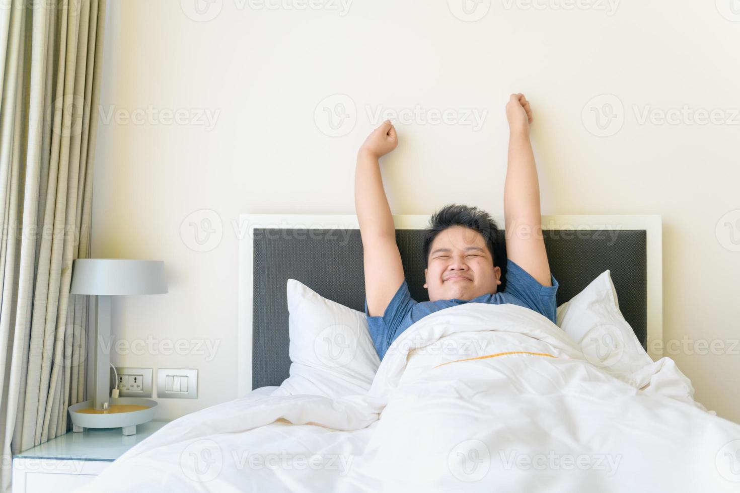 fettleibig Junge aufwachen oben und strecken Arm auf Bett im Morgen. faul Konzept foto