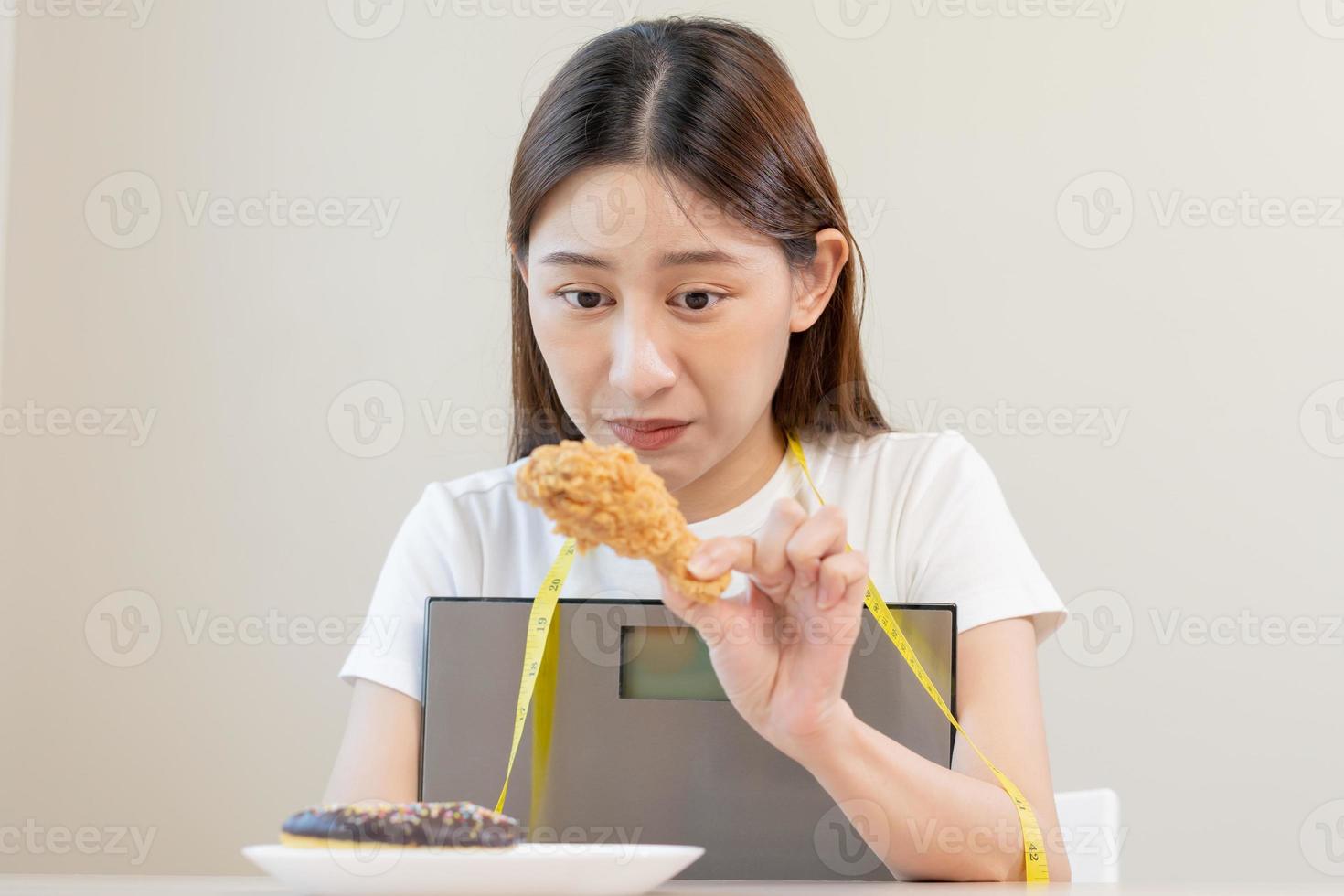 Diät, attraktiv asiatisch jung Frau, Mädchen zurückhaltend zu Essen Krapfen, Bäckerei und gebraten Huhn, schnell Essen zu verlieren, Verlust Gewicht, umarmen Gewicht Waage auf Tabelle beim heim. Hingabe, Versuchung wann hungrig. foto