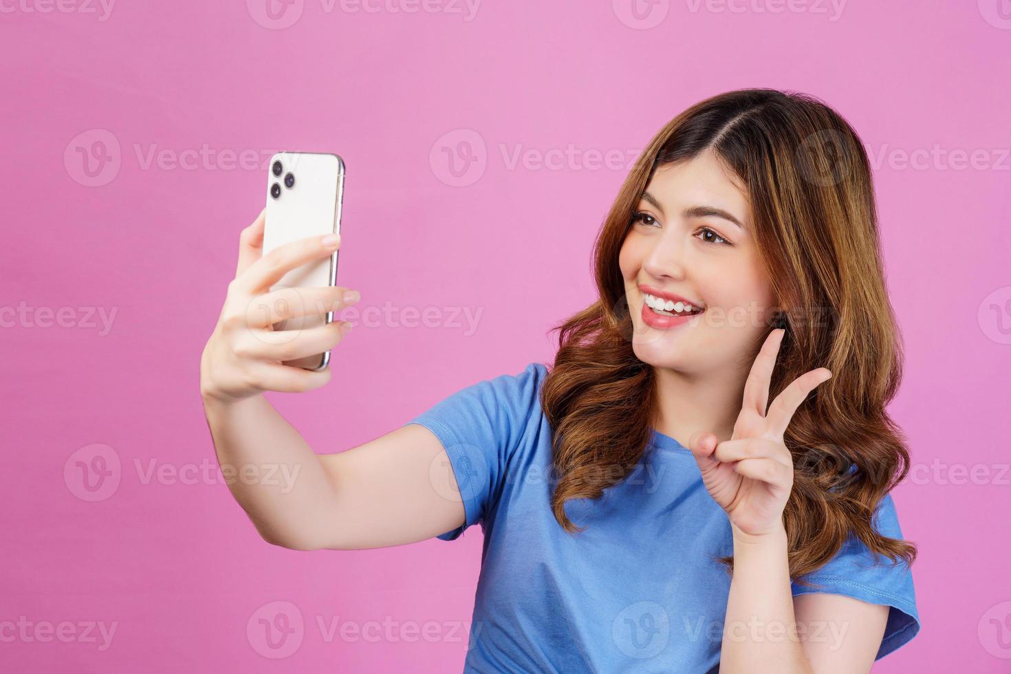 Porträt einer glücklich lächelnden jungen Frau, die ein lässiges T-Shirt-Selfie mit Smartphone trägt, das über rosafarbenem Hintergrund isoliert ist foto