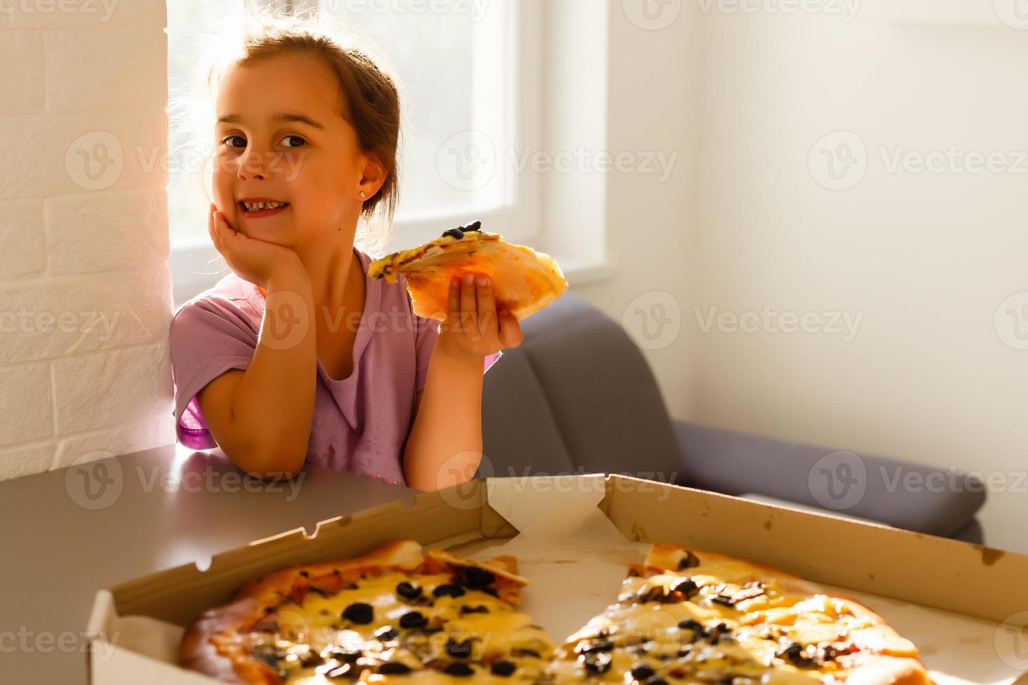 charmant glücklich jung Mädchen Lachen und beißen aus groß Scheibe von frisch gemacht Pizza. sie sitzen beim Weiß Stuhl im Provence Stil Innere, Lächeln und genießen sonnig Tag und lecker Mahlzeit. sie hat lange blond Haar. foto