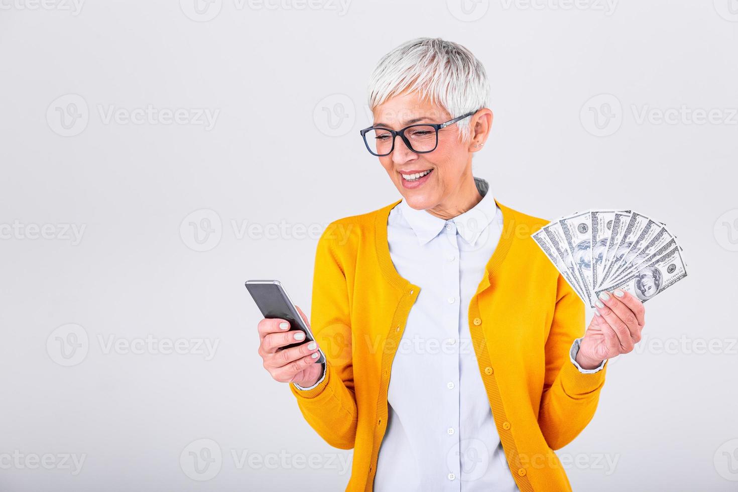 Porträt von ein Erfreut Senior Frau halten Bündel von Geld Banknoten und bezahlen online auf ihr Handy, Mobiltelefon Telefon Über grau Hintergrund foto