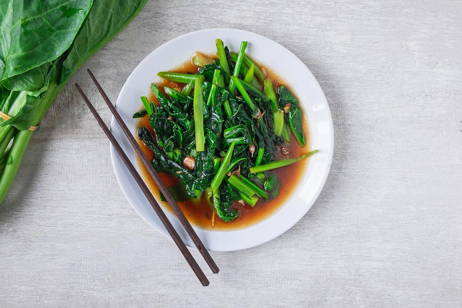 Gebratener chinesischer Grünkohl mit Austernsauce mit Stäbchen auf einem weißen Teller neben frischem chinesischem Grünkohl auf Holztisch foto