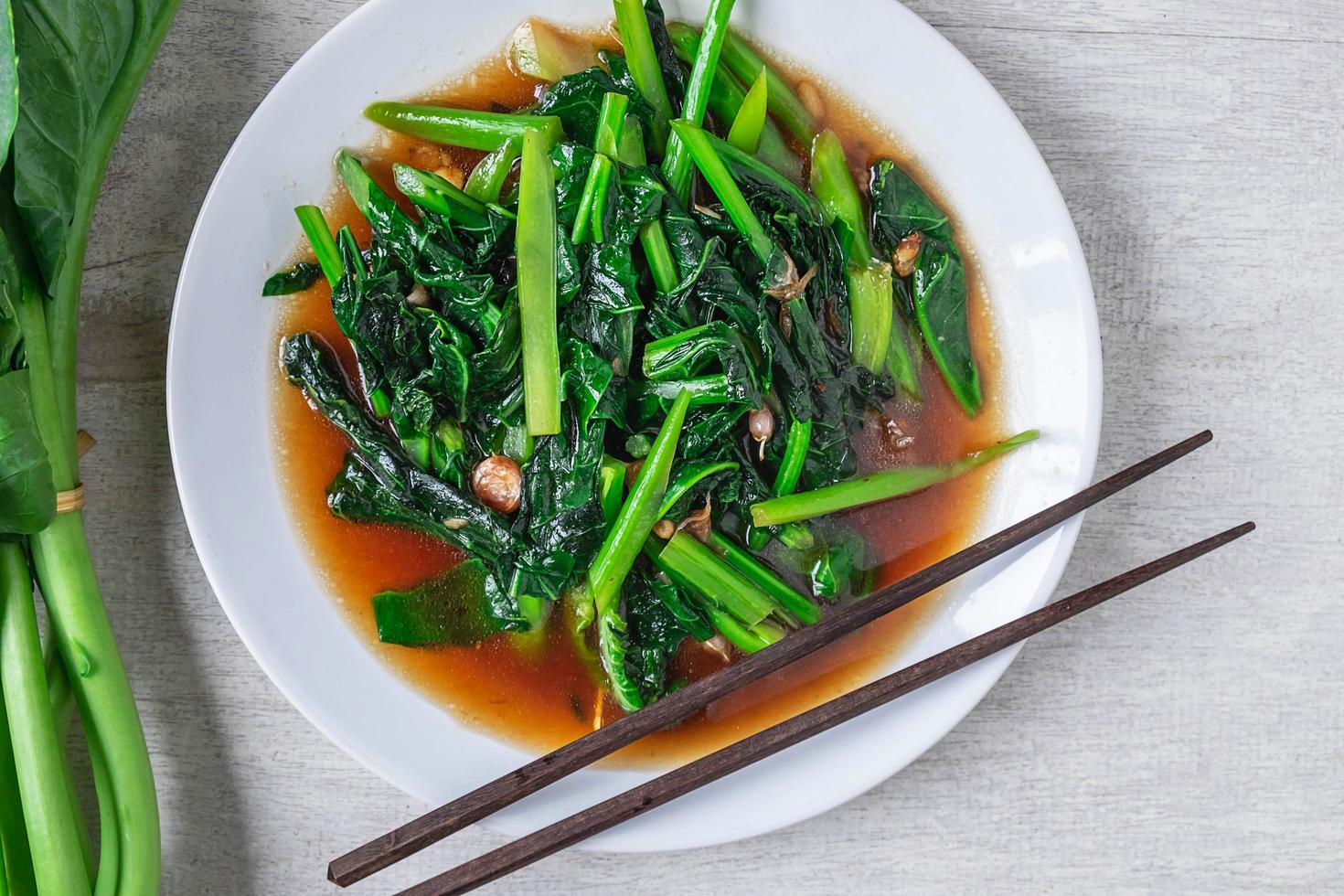 Gebratener chinesischer Grünkohl mit Austernsauce mit Stäbchen neben frischem chinesischem Grünkohl auf einem weißen Teller auf Holztisch foto