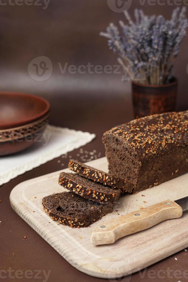 schwarz Brot mit Getreide geschnitten auf ein hölzern Schneiden Tafel gegen ein braun Hintergrund. Nächster zu das Brot Lügen ein Messer, Lehm Platte, Gabel, Löffel und Weiß Baumwolle Tischdecke. Vertikale Schuss foto