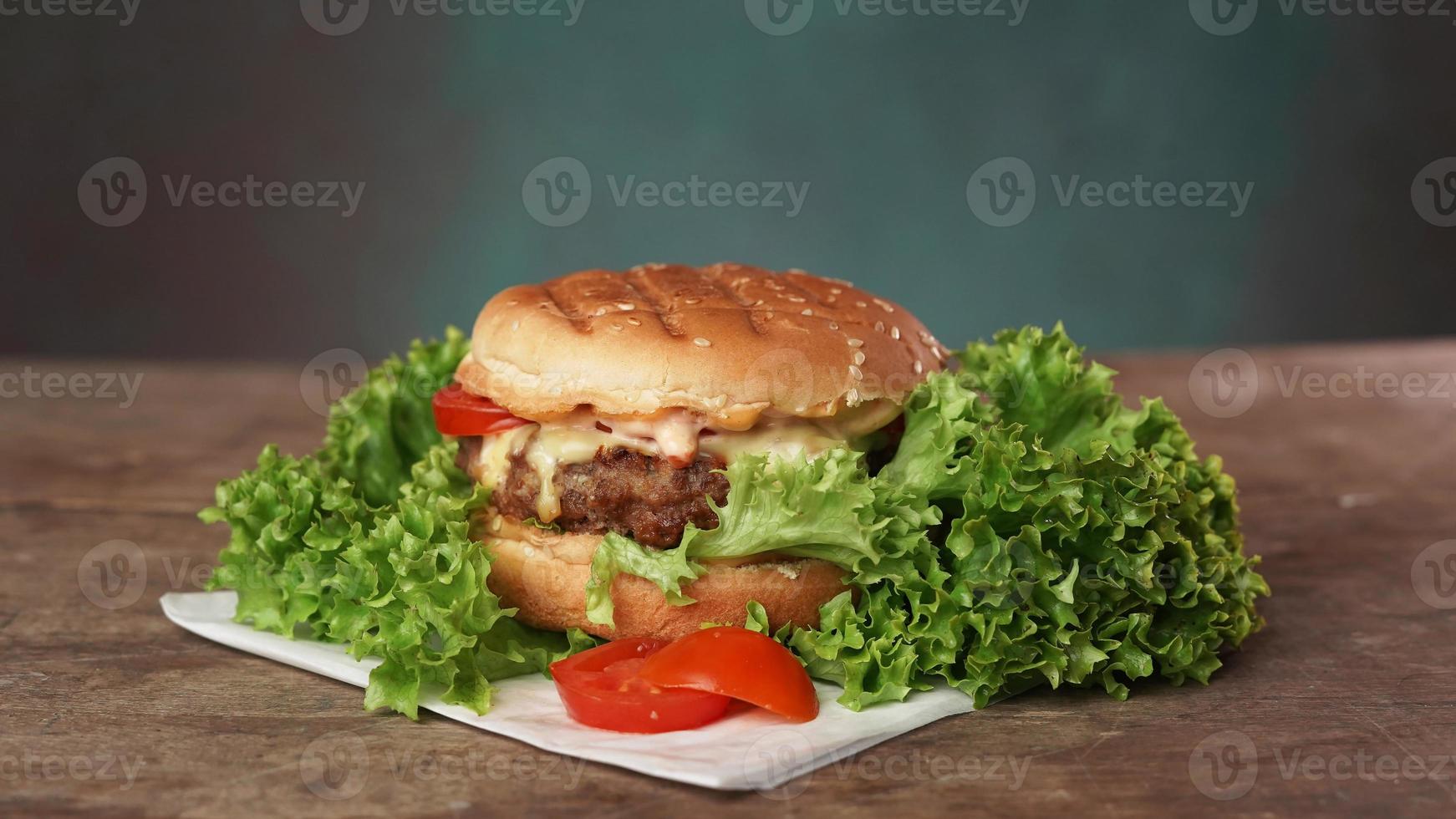 groß lecker Rindfleisch Pastetchen Burger ruht auf Kunst Weiß Papier gegen hölzern Tisch. ein saftig Grün Salat Blatt und ein rot Tomate Lüge in der Nähe von das Burger foto