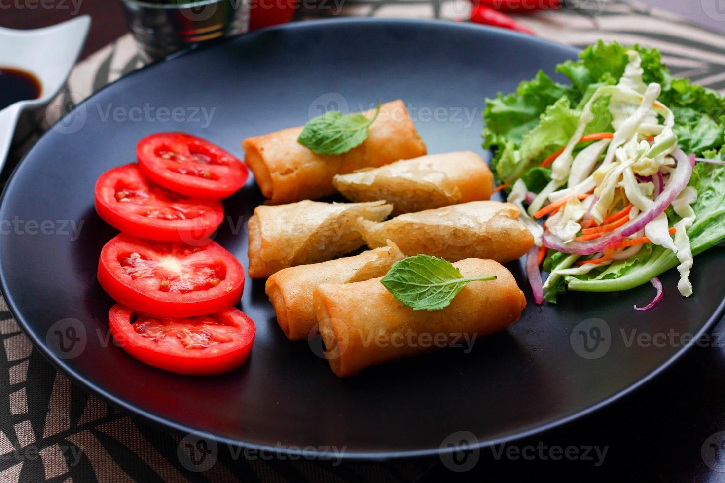 gebraten Frühling Rollen mit Gemüse und Tomaten platziert im ein schwarz Teller auf ein schwarz hölzern Tabelle und Eintauchen Soße. foto