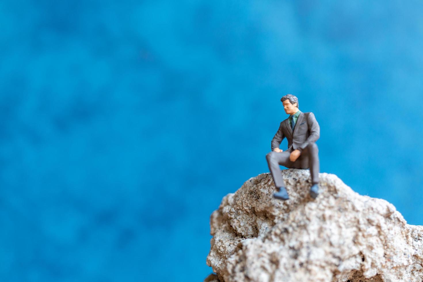 Miniaturgeschäftsmann, der auf einem Felsen mit einem blauen Hintergrund sitzt foto