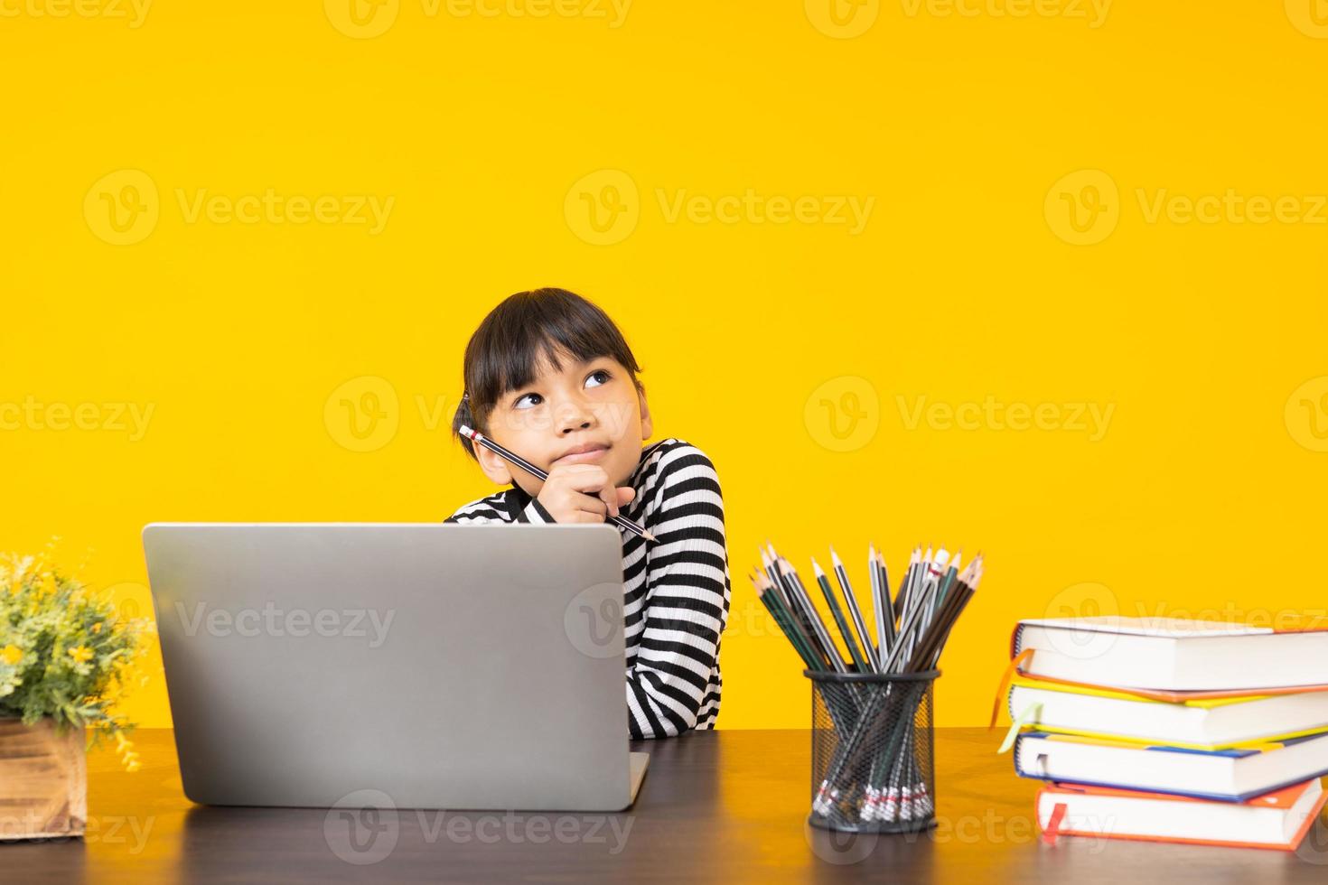asiatisches Mädchen mit denkender Haltung, die mit Laptop, Büchern und Stiften an einem hölzernen Schreibtisch mit gelbem Hintergrund sitzt foto