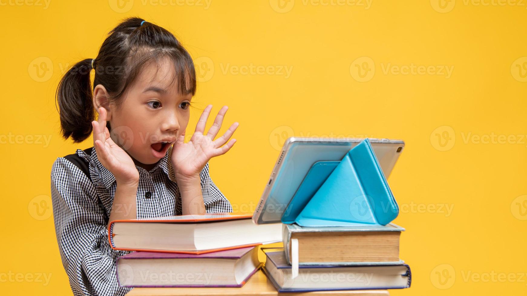 thailändisches Mädchen sieht überrascht aus, wenn es Tablette auf Stapel von Büchern mit gelbem Hintergrund betrachtet foto
