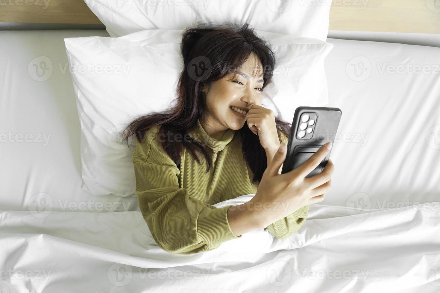 ein lächelnd jung asiatisch Frau ist Lachen während halten ihr Telefon und Lügen auf das Bett foto