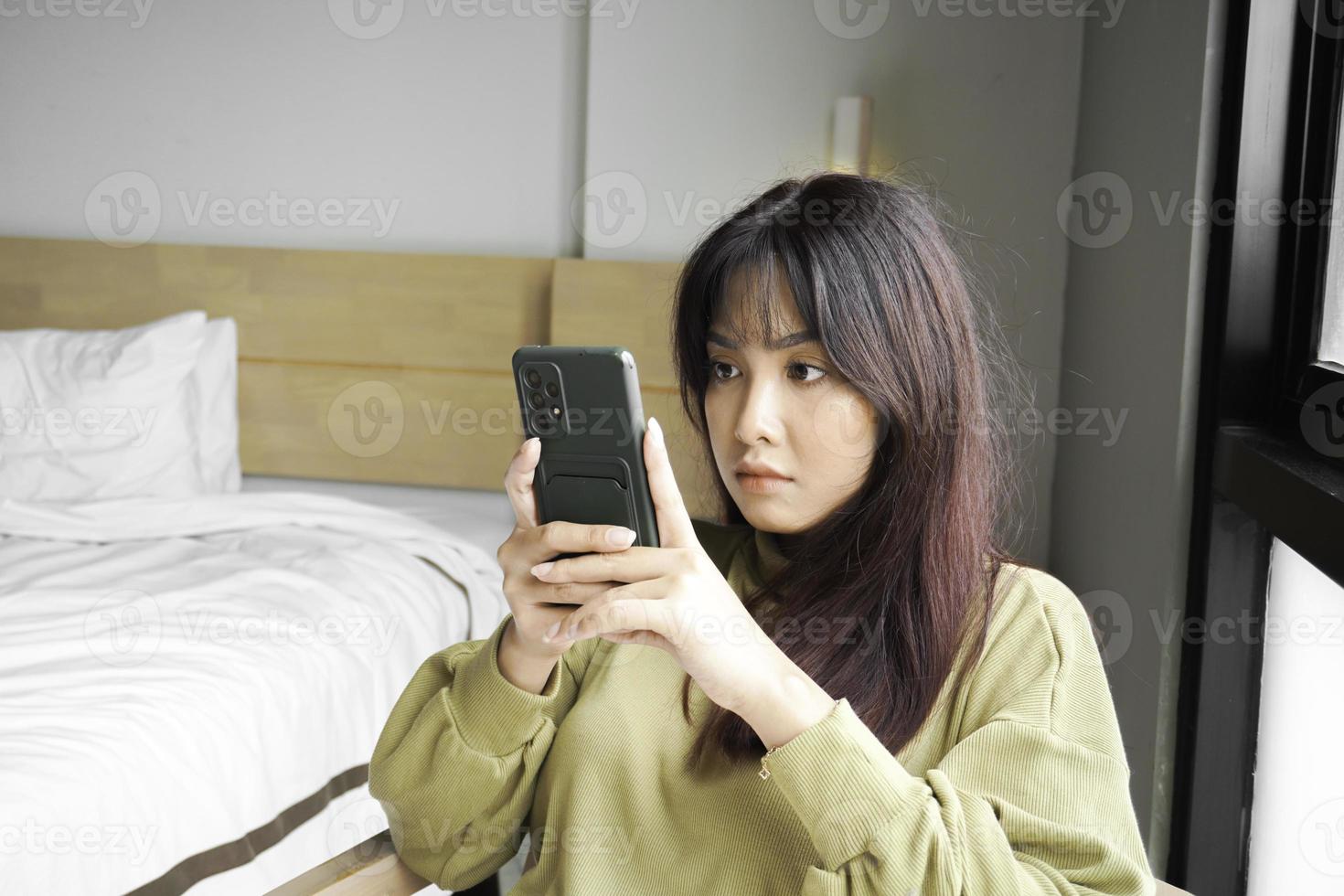 ein jung asiatisch Frau ist Gefühl ernst und konzentriert auf halten ihr Smartphone im Hand während Sitzung im ein Schlafzimmer. foto