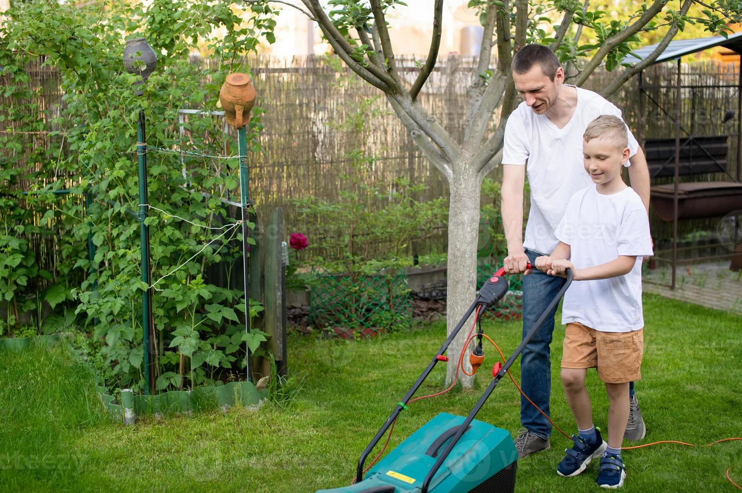 Papa und seine neugierig Sohn mähen das Grün Rasen in der Nähe von das Haus. foto