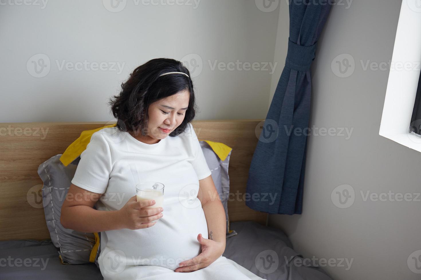 glücklich jung asiatisch schwanger Frau Trinken Milch auf Bett beim Zuhause während halten ihr Bauch und Denken foto