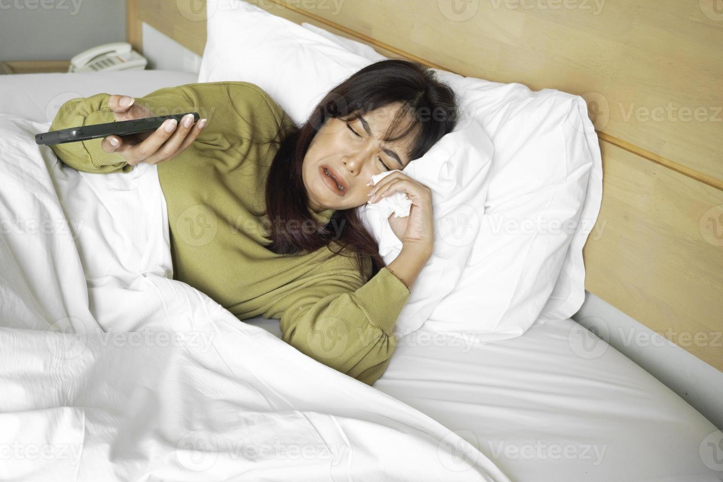 ein traurig jung asiatisch Frau ist Weinen Über ihr Telefon während Lügen auf das Bett foto